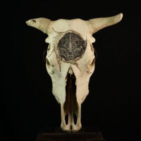 Scultura monumentale realizzata su teschio di Bos taurus, opera manifesto della collezione Exhumed Garden. La composizione si sviluppa attorno al disco frontale lavorato a carving d'alta precisione e rivestito in foglia d’argento, concepito come punto focale di diffrazione della luce per creare un contrasto plastico con le ombre della materia ossea.

L'intervento pittorico combina inchiostro di china e pastello secco sfumato, sovrapposti a stratificazioni d'intaglio diretto che fossilizzano la grafica sulla superficie senza possibilità di ripensamento. Una sintesi di rigore artigianale e tensione materica, pensata per collezioni private d'arte contemporanea.